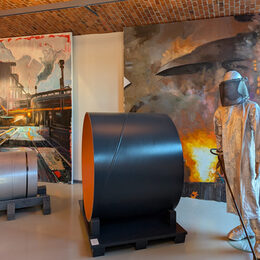 Die beiden Werke von Edward B. Gordon in Hintergrund, davor die Figur eines Probennehmers, ein Stahlrohr-Abschnitt, eine „Coil“ (Bandstahlrolle, Halbfabrikat) und ganz links eine Stahl-Bramme (Vormaterial)