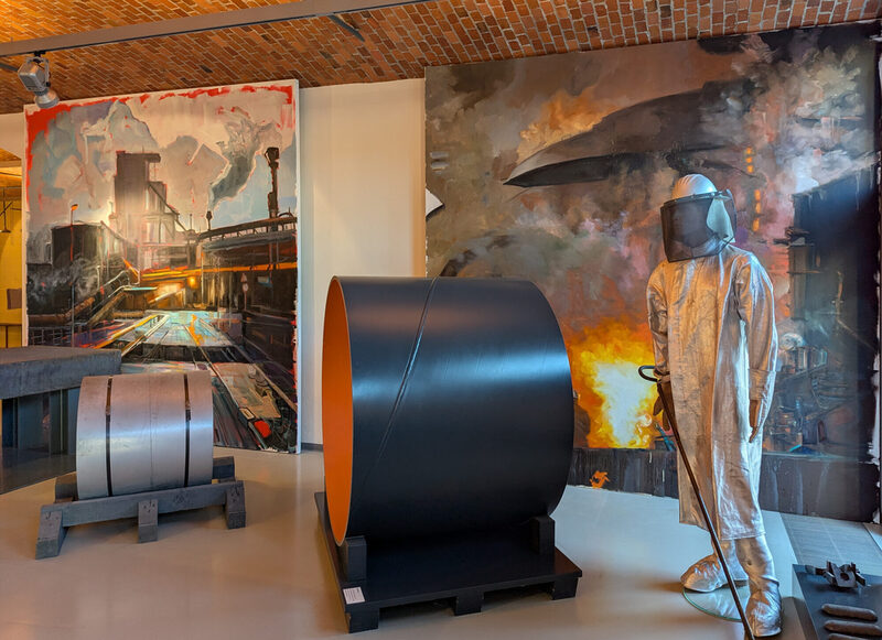 Die beiden Werke von Edward B. Gordon in Hintergrund, davor die Figur eines Probennehmers, ein Stahlrohr-Abschnitt, eine „Coil“ (Bandstahlrolle, Halbfabrikat) und ganz links eine Stahl-Bramme (Vormaterial)