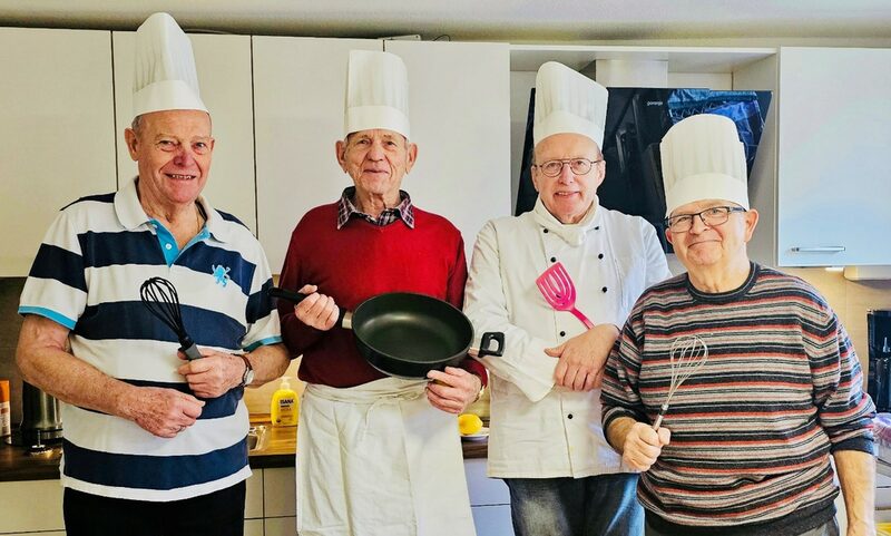 Das Bild zeigt vier ältere Herren mit Kochmützen auf den Köpfen und Kochgeräten in den Händen, die in einer Küche stehen.