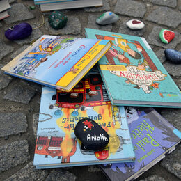 Das Bild zeigt einen Haufen mit Kinderbüchern auf dem Boden. Oben auf liegt ein Stein mit der Aufschrift Antolin.