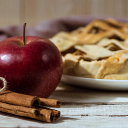 Im Hintergrund ein Apfelkuchen im Vordergrund ein Apfel und Zimtstangen